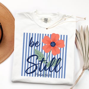 May include: White t-shirt with a floral design and the words "be Still" in blue script. The flower is coral with a blue center. The shirt also has blue and white vertical stripes and the text "Psalm 46:10". A gold necklace with a star pendant is on the shirt.
