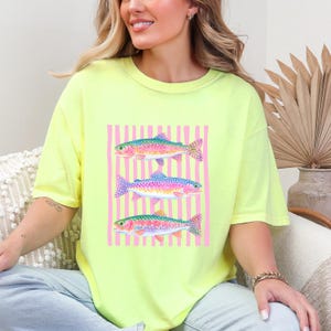 May include: A neon yellow t-shirt featuring a watercolor design of three rainbow trout. The fish are set against a pink striped background. The shirt is a comfortable, oversized fit.