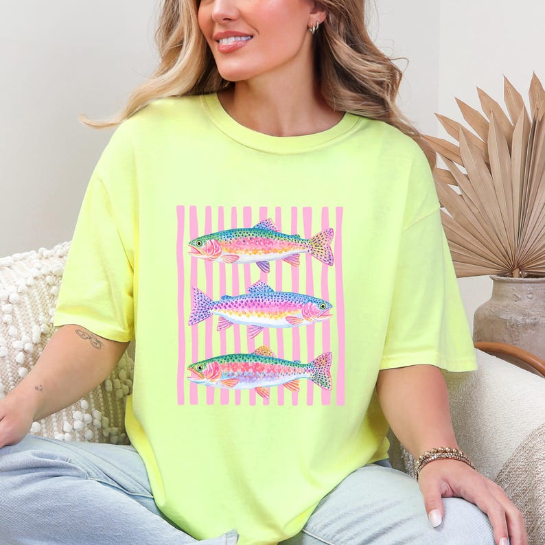 May include: A neon yellow t-shirt featuring a watercolor design of three rainbow trout. The fish are set against a pink striped background. The shirt is a comfortable, oversized fit.
