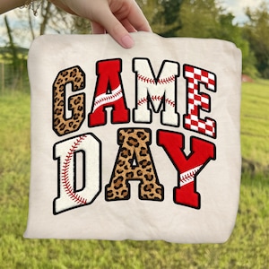 May include: A cream-colored, square pillow with the words "GAME DAY" in bold, block letters. The letters have baseball and leopard print patterns. The baseball letters have red stitching details, and the letter "E" has a red and white checkered pattern.