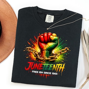 May include: Black t-shirt featuring a fist in the colors of the Pan-African flag, breaking chains, with the word "Juneteenth" and the phrase "Free Ish Since 1865". The shirt has a crew neck and a tag that says "Comfort Colors".