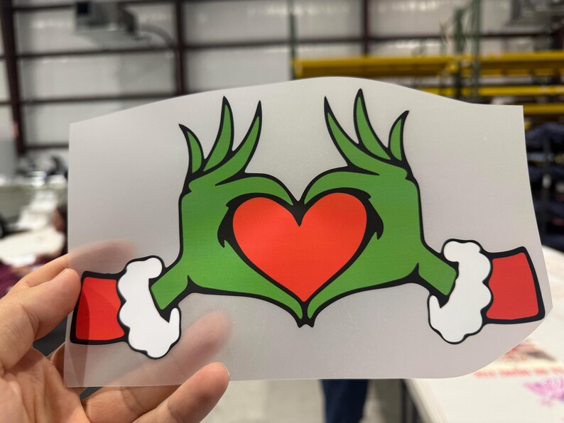 May include: A green hand-shaped design forming a heart with a red center. The hands have white cuffs and red sleeves. The design is on a clear, rectangular sheet.