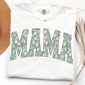 May include: White t-shirt with the word "MAMA" in a floral pattern across the front. The letters are a teal color with white and cream-colored flowers. The shirt has a crew neck and a small tag that says "COMFORT COLORS".