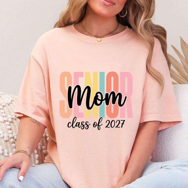 May include: A peach-colored t-shirt featuring the word SENIOR in large, vertical, multi-colored block letters behind the black cursive text Mom and the black printed text class of 2027 centered on the front of the garment.