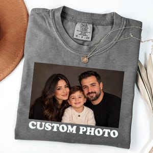 May include: A charcoal gray crew neck t-shirt featuring a rectangular custom photo print of a family of three, consisting of a woman, a young boy, and a man. The text "CUSTOM PHOTO" is printed in white capital letters below the photograph.