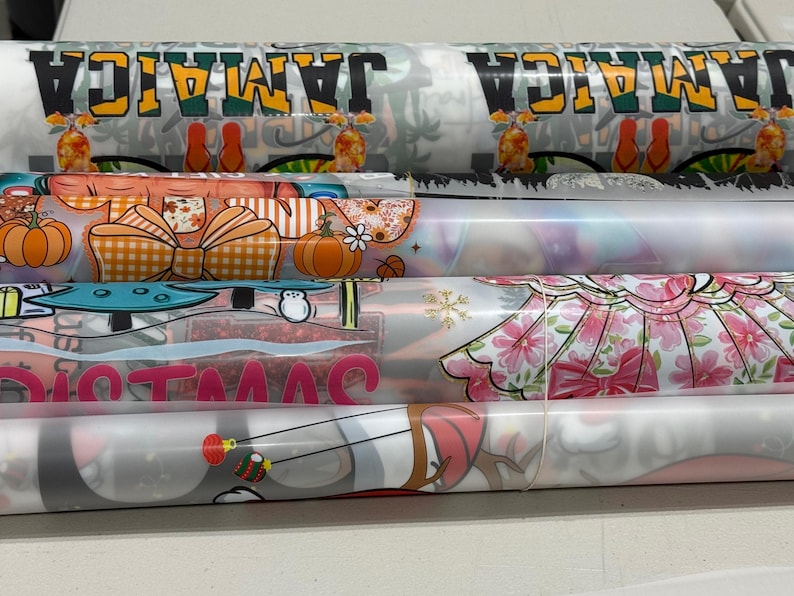 May include: Several rolls of printed fabric are stacked, showcasing various designs. One roll features the word "JAMAICA" in yellow and green. Other rolls display pumpkin, bow, and floral patterns, along with the word "CHRISTMAS".
