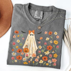 May include: A charcoal gray short-sleeve t-shirt featuring a graphic of a ghost wearing an orange bow and holding a jack-o'-lantern bucket. The ghost is surrounded by colorful wildflowers and small black bats. The shirt has a Comfort Colors label.