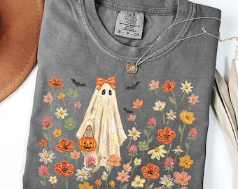 May include: A charcoal gray short-sleeve t-shirt featuring a graphic of a ghost wearing an orange bow and holding a jack-o'-lantern bucket. The ghost is surrounded by colorful wildflowers and small black bats. The shirt has a Comfort Colors label.