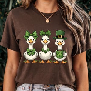 May include: Brown t-shirt featuring three cartoon geese celebrating St. Patrick's Day. The geese are adorned with green bows, shamrocks, and a leprechaun hat, with one holding a mug with a shamrock.