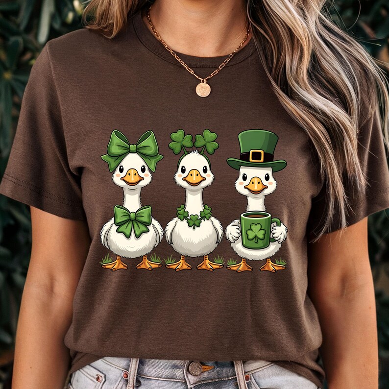 May include: Brown t-shirt featuring three cartoon geese celebrating St. Patrick's Day. The geese are adorned with green bows, shamrocks, and a leprechaun hat, with one holding a mug with a shamrock.