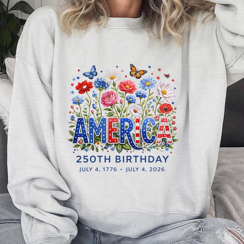 K&ouml;nnte beinhalten: Ein hellgraues Sweatshirt mit einem Blumendesign und dem Wort "AMERICA" in Rot, Wei&szlig; und Blau. Das Design umfasst Schmetterlinge, Blumen und den Text "250TH BIRTHDAY JULY 4, 1776 &bull; JULY 4, 2026".