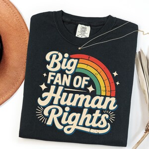 May include: Black t-shirt with the text "Big Fan of Human Rights" in a retro font with a rainbow design. A gold necklace with a star pendant is on the shirt. A brown hat is in the upper left corner.