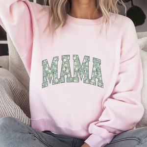 May include: A light pink sweatshirt with the word "MAMA" in large, floral-patterned letters across the front. The floral pattern features a light blue-green background with small white flowers. The sweatshirt has a crew neck and long sleeves.