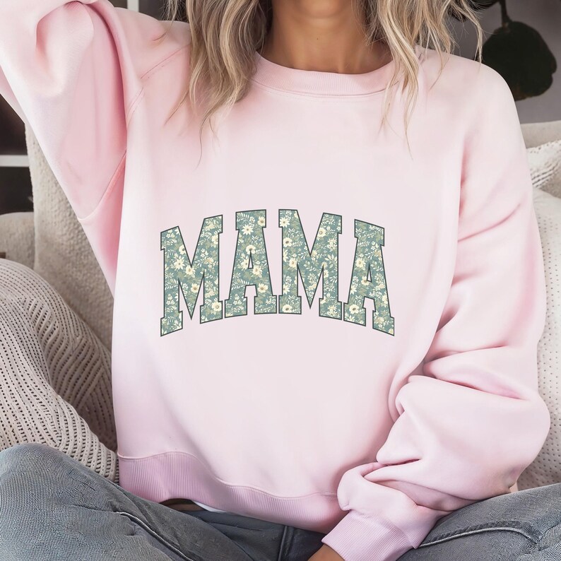 May include: A light pink sweatshirt with the word "MAMA" in large, floral-patterned letters across the front. The floral pattern features a light blue-green background with small white flowers. The sweatshirt has a crew neck and long sleeves.