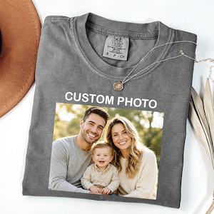 May include: A charcoal gray crew neck t-shirt featuring a rectangular graphic print of a family portrait with a man, a woman, and a baby. The text "CUSTOM PHOTO" is printed in white above the image. The shirt has a Comfort Colors brand label.