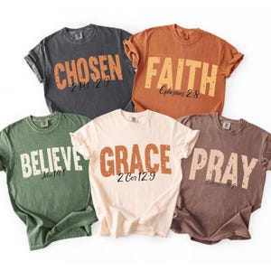 May include: Five short-sleeve crew neck t-shirts in charcoal, orange, olive green, cream, and brown. Each shirt features a large, distressed-style word printed in the center: CHOSEN, FAITH, BELIEVE, GRACE, or PRAY, with a corresponding Bible verse below.