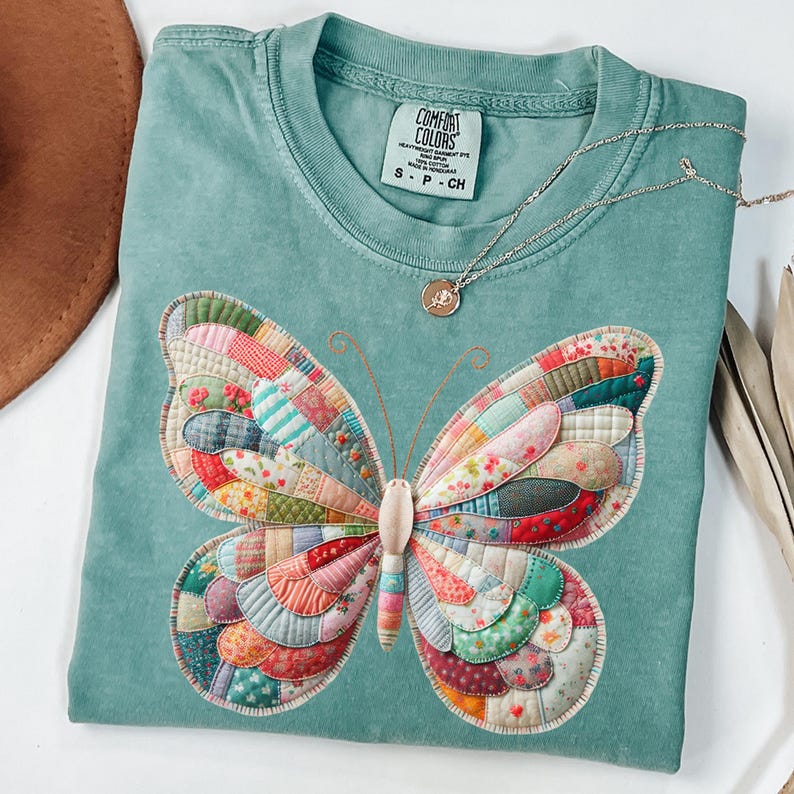 Puede incluir: Una camiseta Comfort Colors de color verde azulado con un gran aplique de mariposa colorido. Las alas de la mariposa est&aacute;n hechas de tela de retazos con varios patrones florales y geom&eacute;tricos. Un collar plateado con un peque&ntilde;o colgante descansa sobre la camisa.