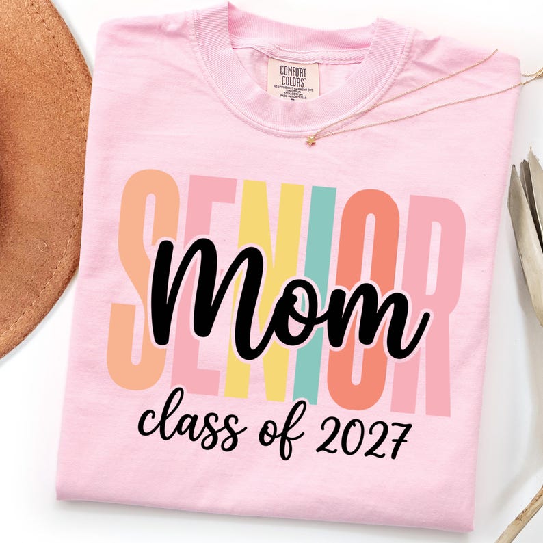 May include: A light pink short-sleeve t-shirt featuring the word SENIOR in large, vertical, multi-colored block letters. Overlaid in black script is the word Mom, followed by the text class of 2027 in a smaller black cursive font below.