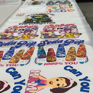 May include: Printed fabric with diverse designs, including text and illustrations. The designs feature phrases like "Everything's Fine" and "Real Thick Face." Other graphics include cartoon characters and soda bottle illustrations.