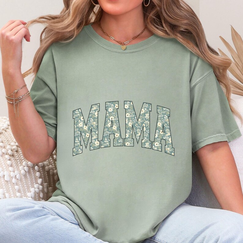 May include: Sage green t-shirt with the word "MAMA" in a floral print design. The letters are outlined in a darker green, and the floral pattern features small white flowers. The shirt has short sleeves and a relaxed fit.