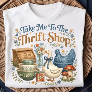 May include: White t-shirt with a colorful graphic design featuring a goose, chicken, mushrooms, bowls, and a bread box. The text "Take Me To The Thrift Shop" is displayed in a decorative font. The design is surrounded by floral elements.