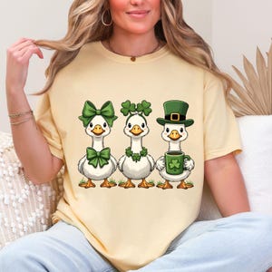 May include: A light yellow t-shirt featuring three cartoon geese. Each goose is decorated with St. Patrick's Day themed accessories, including green bows, shamrock garlands, and a green hat. One goose holds a mug with a shamrock.
