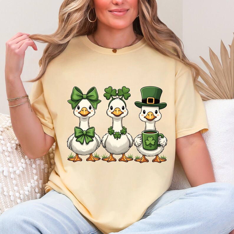 May include: A light yellow t-shirt featuring three cartoon geese. Each goose is decorated with St. Patrick's Day themed accessories, including green bows, shamrock garlands, and a green hat. One goose holds a mug with a shamrock.