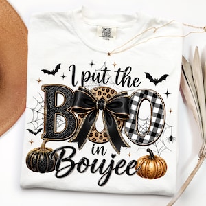 May include: A white t-shirt featuring the text I put the BOO in Boujee. The design includes a black glitter letter B, a leopard print letter O, and a black and white buffalo plaid letter O with a large black bow. Accents include pumpkins, bats, and spiderwebs.