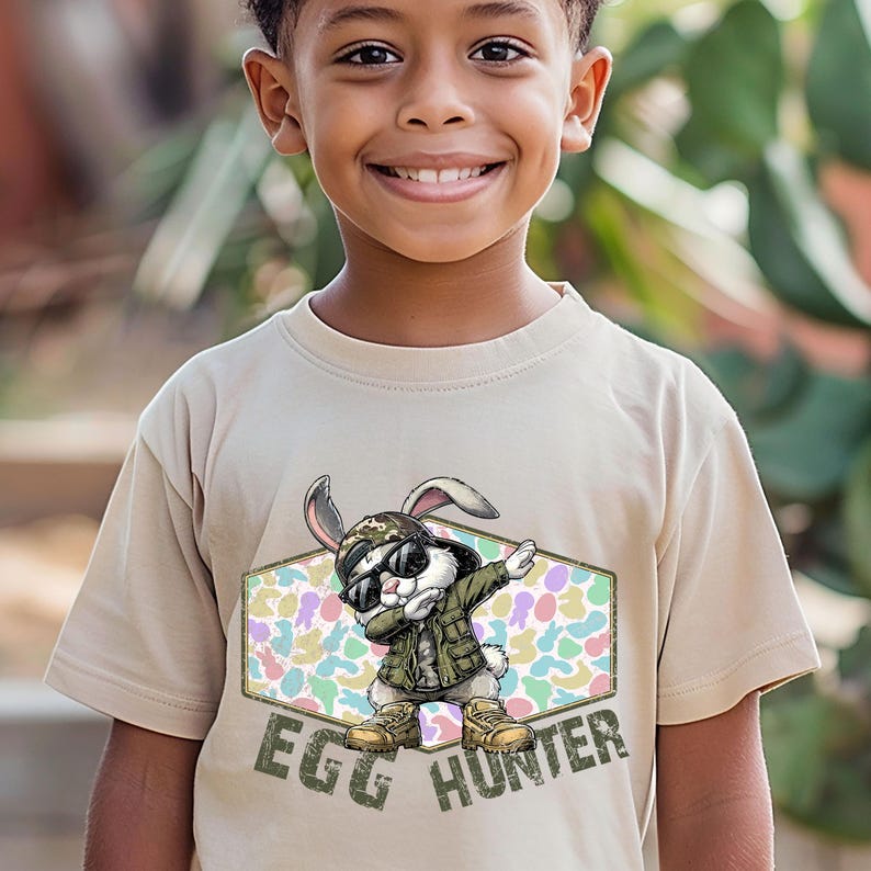 May include: Tan t-shirt featuring a cartoon rabbit wearing sunglasses, a camouflage jacket, and boots, doing the dab. The text "EGG HUNTER" is printed below the rabbit in green letters. The background has a pastel egg pattern.