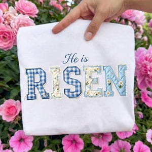 May include: White sweatshirt with the embroidered phrase "He is RISEN". The letters are in a patchwork style, with blue gingham and floral patterns. The sweatshirt is held up by a hand, with pink flowers in the background.