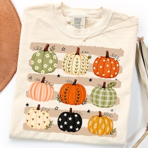 May include: A cream-colored t-shirt featuring a graphic print of nine pumpkins arranged in three rows. The pumpkins display various patterns including daisies, polka dots, plaid, hearts, stars, and vines, interspersed with small black stars.