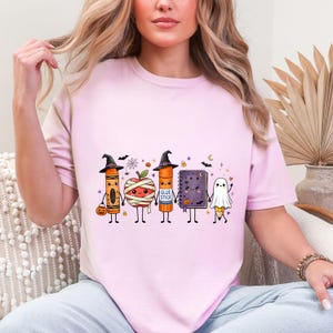 May include: A light pink t-shirt featuring a graphic of five cute, anthropomorphic school supplies dressed for Halloween: a crayon witch, a mummy apple, a glue stick witch, a purple notebook, and a ghost pencil holding hands.