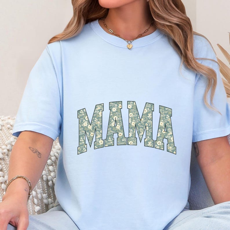 May include: Light blue t-shirt with the word "MAMA" in a floral design. The letters are filled with a pattern of small white and green flowers. The shirt has short sleeves and a crew neck.