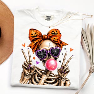 May include: A white t-shirt featuring a graphic of a skeleton wearing heart-shaped sunglasses with spiderweb patterns, a large orange bow with jack-o'-lantern faces, and blowing a pink bubblegum bubble while making peace signs with both hands.