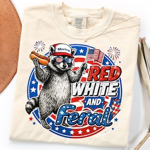 May include: Cream-colored t-shirt featuring a raccoon wearing sunglasses and a hat with the word "'Merica". The raccoon holds a hot dog and an American flag. The shirt has the words "Red White and Feral".