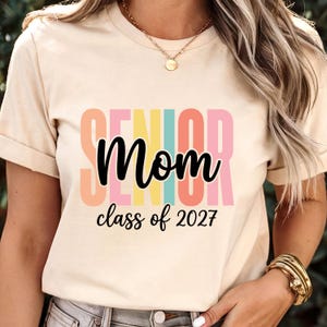 May include: A cream-colored short-sleeve t-shirt featuring the word SENIOR in large, multicolored block letters behind the black cursive text Mom and the black printed text class of 2027. The shirt has a crew neckline and a standard fit.