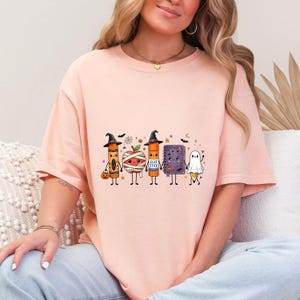May include: A light peach-colored t-shirt featuring a graphic print of five cartoon school supplies dressed for Halloween: a crayon, a mummy apple, a glue stick, a purple notebook, and a pencil ghost, all holding hands with small bats and stars.