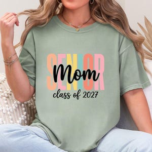 May include: A sage green short-sleeve t-shirt featuring the text "SENIOR" in large, vertical, multi-colored block letters behind the word "Mom" in black cursive script, with the text "class of 2027" printed in black below the main design.