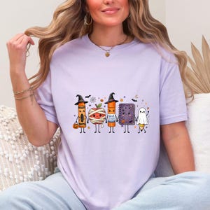 May include: A light purple t-shirt featuring a graphic of five anthropomorphic school supplies dressed for Halloween, including a crayon, a mummy apple, a glue stick, a notebook, and a ghost pencil, all holding hands with small bats and stars.