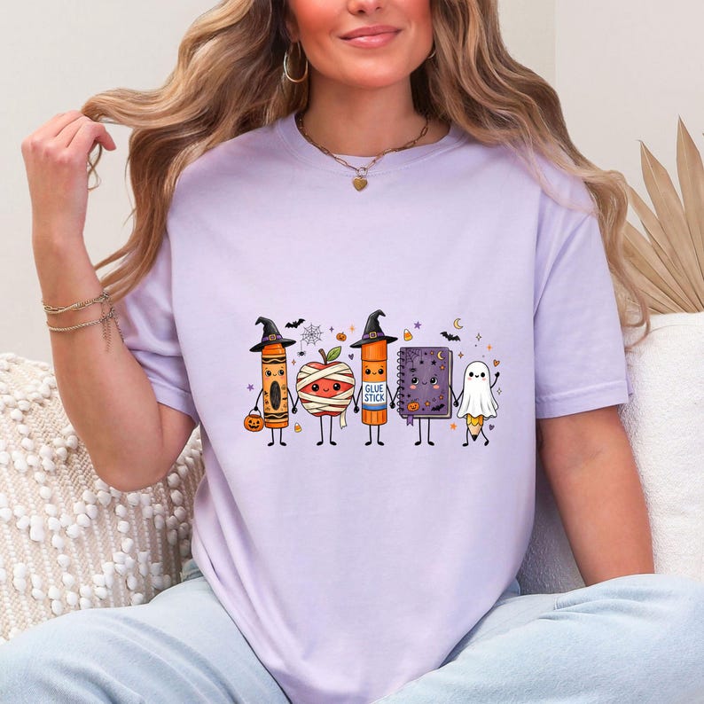 May include: A light purple t-shirt featuring a graphic of five anthropomorphic school supplies dressed for Halloween, including a crayon, a mummy apple, a glue stick, a notebook, and a ghost pencil, all holding hands with small bats and stars.
