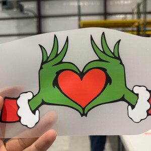 May include: A green hand-shaped heart design with a red heart in the center. The hands have green fingers and red cuffs with white trim. The design is on a clear, rectangular background.