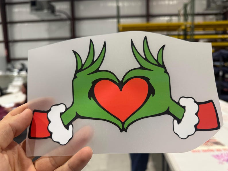 May include: A green hand-shaped heart design with a red heart in the center. The hands have green fingers and red cuffs with white trim. The design is on a clear, rectangular background.