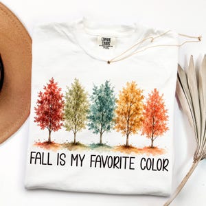 May include: A white short-sleeve t-shirt featuring a graphic of five watercolor-style trees in shades of red, green, teal, yellow, and orange. Below the trees, the text reads "FALL IS MY FAVORITE COLOR" in a bold, black, sans-serif font.