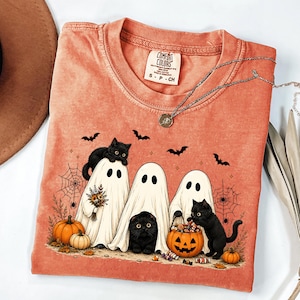 May include: A coral-colored t-shirt with a graphic print featuring three white ghosts and two black cats. The design includes pumpkins, spiderwebs, and bats. The shirt has a Comfort Colors label at the neck, which reads Comfort Colors, S-P-CH.