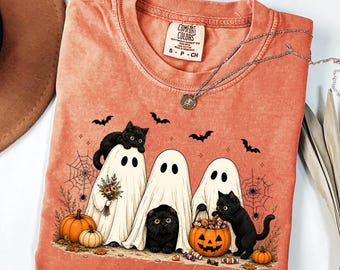 May include: A coral-colored t-shirt with a graphic print featuring three white ghosts and two black cats. The design includes pumpkins, spiderwebs, and bats. The shirt has a Comfort Colors label at the neck, which reads Comfort Colors, S-P-CH.