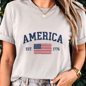 May include: A light grey short-sleeved t-shirt featuring the dark blue text AMERICA in an arched font above a distressed graphic of the United States flag and the text EST. 1776 in dark blue, with a gold chain necklace worn around the neck.