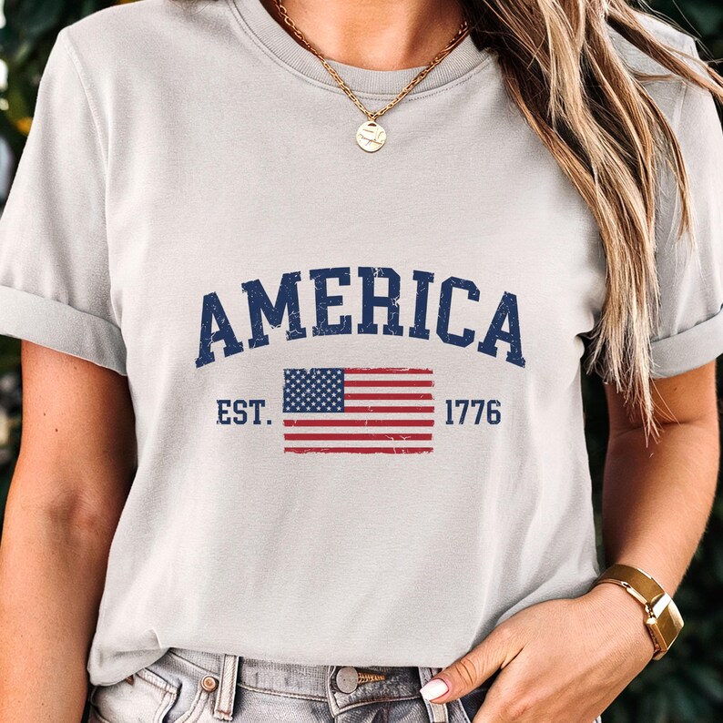 May include: A light grey short-sleeved t-shirt featuring the dark blue text AMERICA in an arched font above a distressed graphic of the United States flag and the text EST. 1776 in dark blue, with a gold chain necklace worn around the neck.