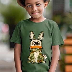 Puede incluir: Camiseta verde con un dise&ntilde;o de conejo de dibujos animados. El conejo lleva una gorra de camuflaje con el texto "EGG HUNTER". El conejo tiene un cuerpo de camuflaje y una cola blanca y esponjosa. La camiseta es de color verde liso.