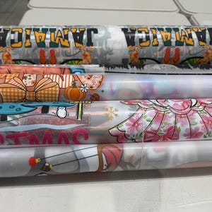 May include: Several rolled-up printed designs are visible, including one with "JAMAICA" in green, yellow, and black. Other designs feature pumpkins, bows, mushrooms, and floral patterns. The rolls are likely for crafting or DIY projects.
