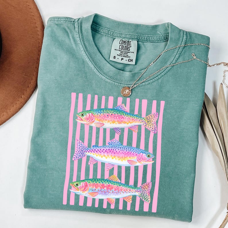 May include: A seafoam green t-shirt featuring a graphic of three colorful rainbow trout set against a pink striped background. The shirt has a crew neck and short sleeves. A silver necklace with a pendant is draped across the shirt.